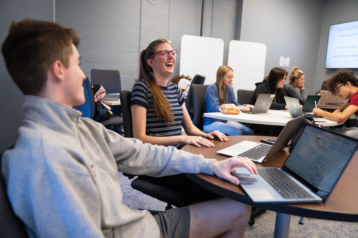 学生拿着笔记本电脑对着桌子笑. 更多的学生在后台和前台.